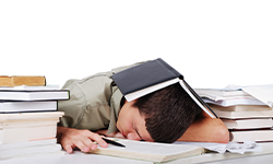 Male student sleeping with pile of books around him and open book over his head