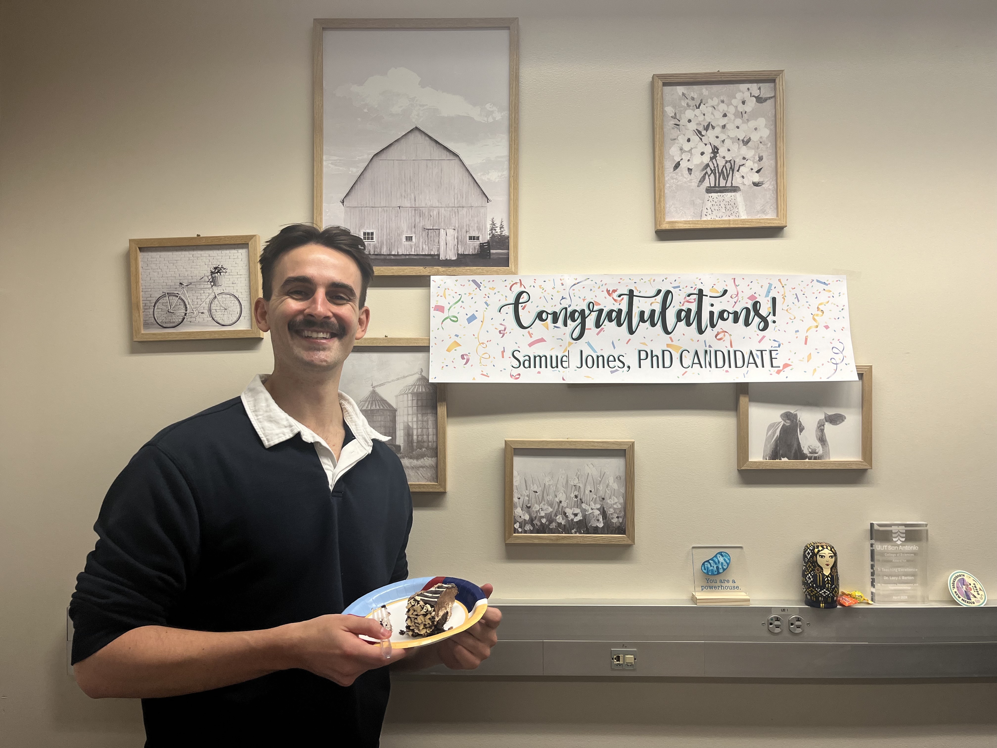 Ph.D. Candidate Samuel Jones standing in front of a "congratulations" sign that's hanging on a wall with various framed images, holding a piece of cake, smiling at camera
