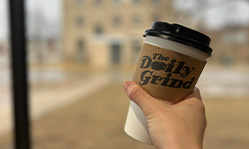 Closeup of hand holding a hot coffee in a to-go cup with “Daily Grind” logo on a cup