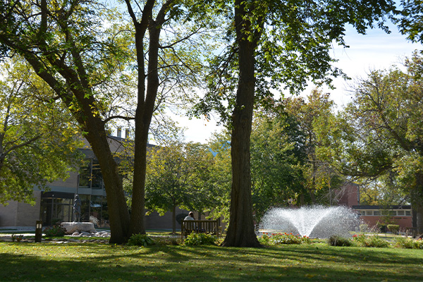 Outdoor photo of Lund Pond with fountain running in the background and a student sitting on a bench by the pond
