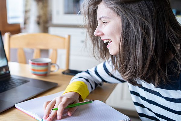 Female with dark hair and wearing a black and white striped shirt smiling at the computer with pen in hand and graph paper on table